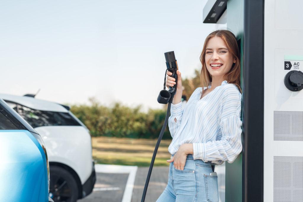 Junge Frau lehnt an einer Wallbox mit einem Ladekabel für Elektroautos in der Hand und lächelt